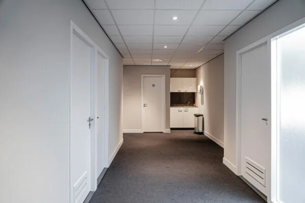 Witte, strak afgewerkte gang met meerdere deuren en een kitchenette aan het einde, in een pand aan de Burgemeester van Karnebeeklaan.